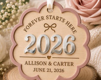 Op de afbeelding: Een geschulpt houten ornament met een oudroze rand en een wit lint. De tekst luidt FOREVER STARTS HERE, 2026, ALLISON & CARTER en JUNE 21, 2026. Het bevat een lasergegraveerde strik, een klein hartje en verhoogde zilveren 2026-cijfers.