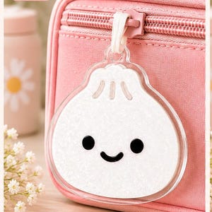 May include: A dumpling-shaped lunch bag tag with a white glittery front featuring a black smiling face and a light wood back engraved with the text this lunch belongs to AMY and a heart symbol, attached to a pink zippered lunch bag.
