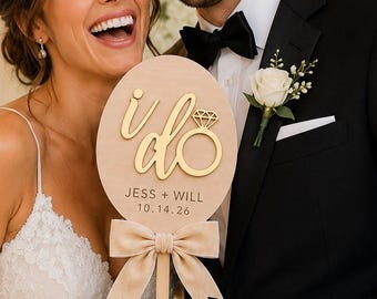 Op de afbeelding: Een rond houten trouwbord met de tekst "i do" en een icoon van een diamanten ring, gepersonaliseerd met de namen "JESS + WILL" en de datum "10.14.26". Het bord is bevestigd aan een houten handvat en versierd met een beige fluwelen strik.