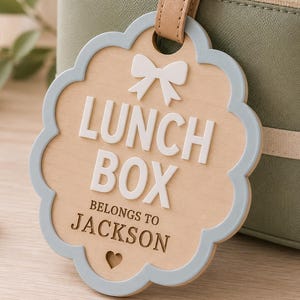 May include: A scalloped wooden tag with a light blue border, featuring a white bow icon, the text LUNCH BOX, the text BELONGS TO JACKSON, and a small heart cutout. The tag is attached to a sage green lunch bag with a tan leather-like strap.