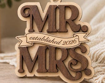 Op de afbeelding: Een gelaagd houten ornament met donkerbruine uitgesneden letters die MR. en MRS. vormen op een lichte houten achtergrond. Een centrale banner is gegraveerd met de tekst established 2026. Het ornament hangt aan een crèmekleurig satijnen lint.