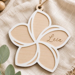May include: A five-petaled flower-shaped wooden ornament with a white border and a natural light wood centre. The name Leia is engraved in cursive script on one of the petals. A small hole at the top holds a thin brown twine string for hanging.