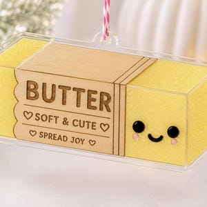 Puede incluir: Un adorno rectangular con forma de mantequilla encerrado en acrílico transparente. El centro presenta un panel de madera clara grabado con el texto BUTTER, SOFT & CUTE y SPREAD JOY. Los extremos son amarillos con una cara sonriente negra.