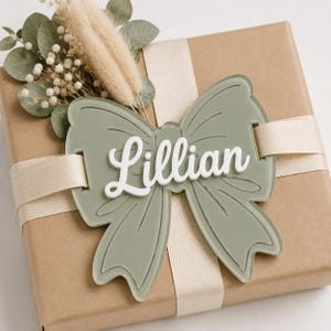Puede incluir: Una etiqueta de regalo con forma de lazo de acrílico verde salvia con el nombre Lillian en letras blancas en relieve. El lazo está sujeto a una caja de regalo envuelta en papel marrón con una cinta de satén crema y eucalipto seco.