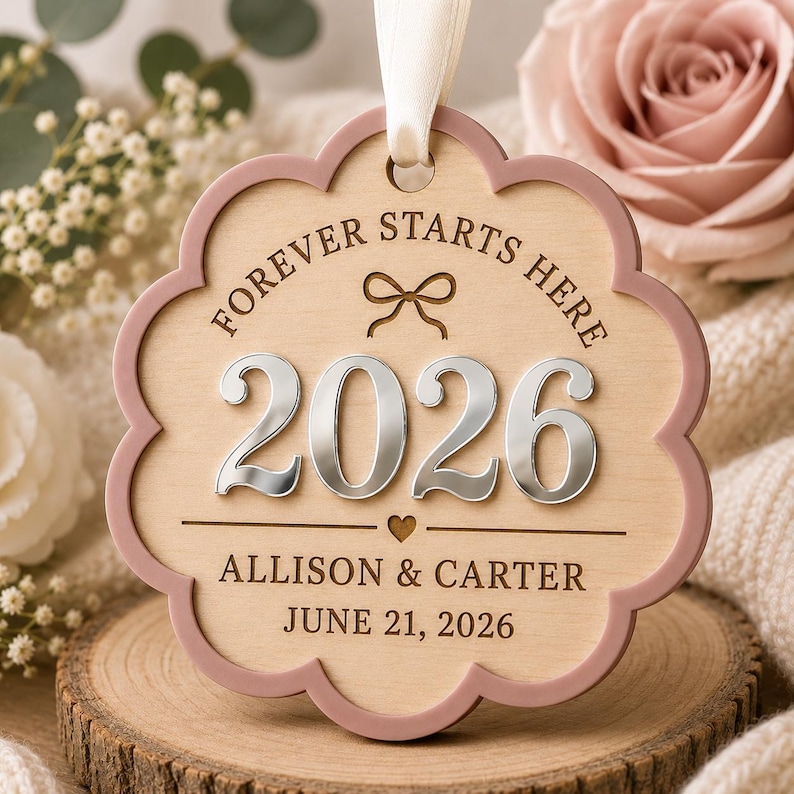 Op de afbeelding: Een geschulpt houten ornament met een oudroze rand en een wit lint. De tekst luidt FOREVER STARTS HERE, 2026, ALLISON & CARTER en JUNE 21, 2026. Het bevat een lasergegraveerde strik, een klein hartje en verhoogde zilveren 2026-cijfers.