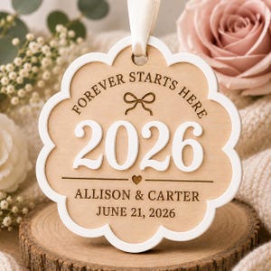 Op de afbeelding: Een geschulpt houten ornament met een witte rand en een cr&egrave;mekleurig lint. De tekst luidt FOREVER STARTS HERE, 2026, ALLISON & CARTER en JUNE 21, 2026. Het heeft een klein gegraveerd strik- en hartsymbool op het lichte houten oppervlak.