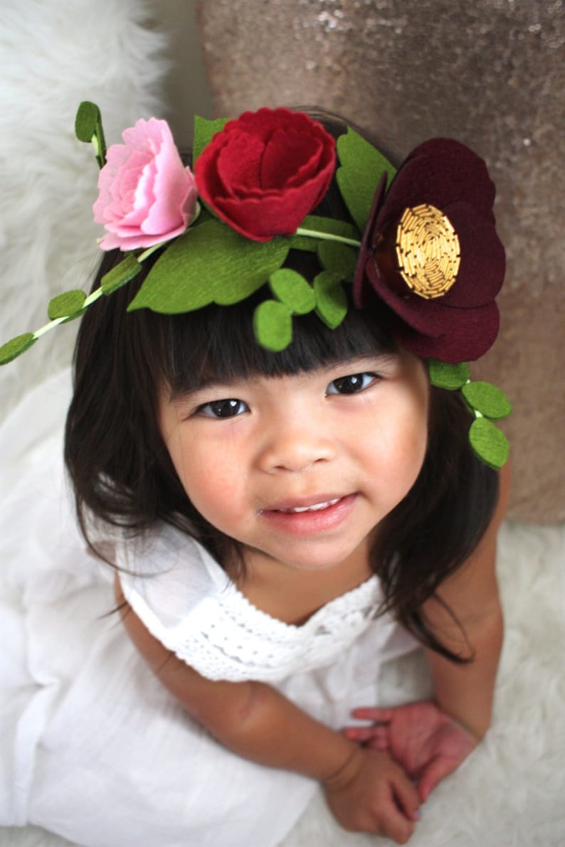 May include: A floral crown featuring felt flowers in shades of light pink, deep red, and dark burgundy. The arrangement includes green felt leaves and stems, with the dark burgundy flower accented by a circular center of gold metallic beads.