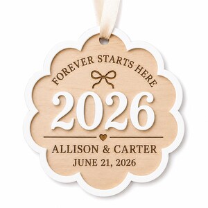 Op de afbeelding: Een geschulpt ornament van licht hout met een witte rand en een cr&egrave;mekleurig lint. Het bevat de gegraveerde tekst "FOREVER STARTS HERE", een strikicoon, het witte verhoogde jaartal "2026", een klein hartje en de namen "ALLISON & CARTER" met "JUNE 21, 2026".