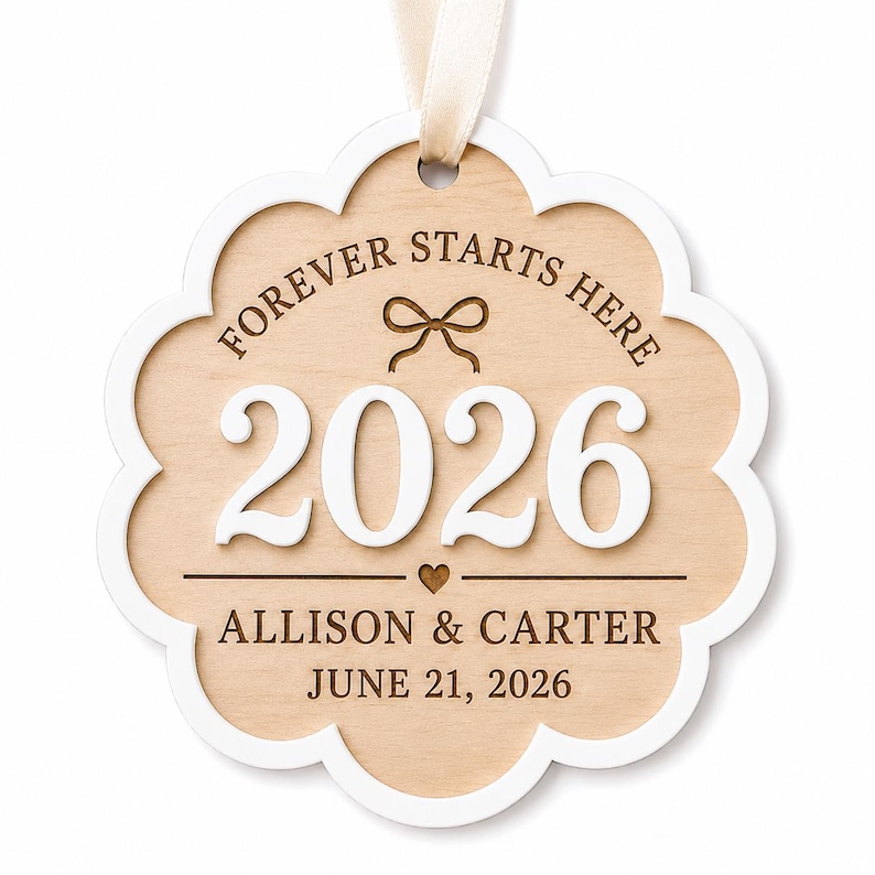 Op de afbeelding: Een geschulpt ornament van licht hout met een witte rand en een cr&egrave;mekleurig lint. Het bevat de gegraveerde tekst "FOREVER STARTS HERE", een strikicoon, het witte verhoogde jaartal "2026", een klein hartje en de namen "ALLISON & CARTER" met "JUNE 21, 2026".