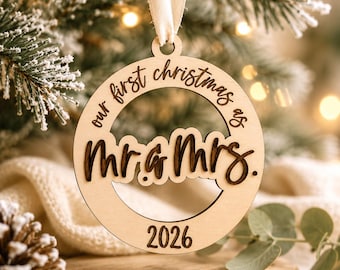 Op de afbeelding: Een ronde kerstversiering van lichtgekleurd hout met een crèmekleurig lint aan de bovenkant. De versiering is gegraveerd met de tekst "our first christmas as" en "2026", met "Mr & Mrs." in donkere, verhoogde cursieve letters.