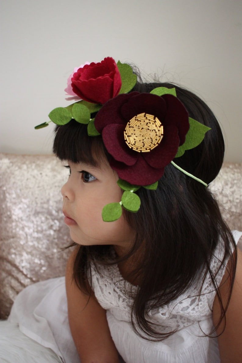 May include: A headband featuring felt flowers in shades of deep burgundy, red, and light pink, accented with green felt leaves. The center of the largest burgundy flower is embellished with gold-toned cylindrical beads arranged in a circular pattern.