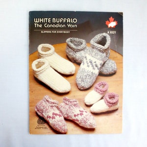 Peut inclure: Une brochure de modèles de tricot pour des pantoufles, avec un logo de bison blanc et le texte "White Buffalo The Canadian Yarn Slippers for Everybody" et "A 6521". La brochure montre des images de pantoufles tricotées en blanc, gris et rose.