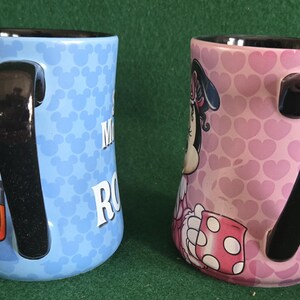 May include: Two ceramic mugs with black handles and interiors. The left mug is light blue with a repeating Mickey Mouse head pattern and a partial graphic. The right mug is pink with a repeating heart pattern and a partial Minnie Mouse graphic.