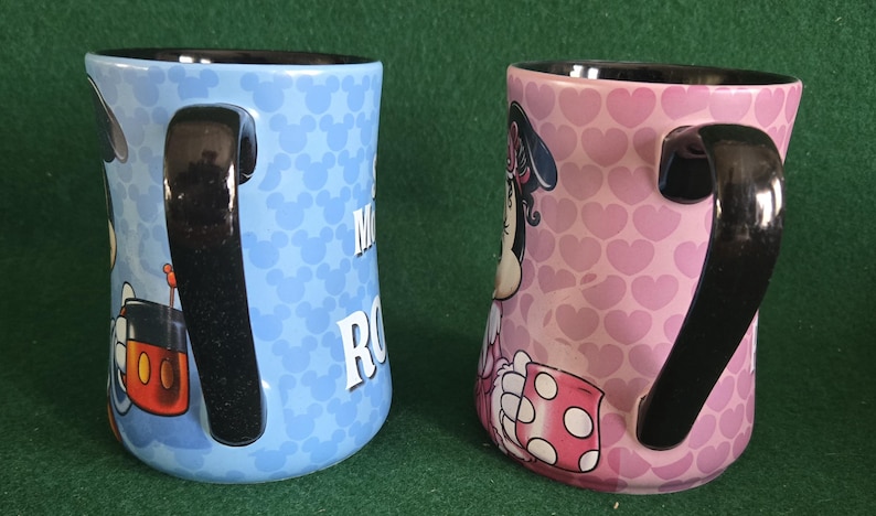 May include: Two ceramic mugs with black handles and interiors. The left mug is light blue with a repeating Mickey Mouse head pattern and a partial graphic. The right mug is pink with a repeating heart pattern and a partial Minnie Mouse graphic.