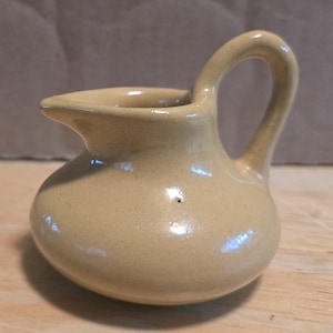 May include: A small, light tan ceramic creamer pitcher with a rounded, bulbous body, a short pouring spout, and a single curved handle attached to the side. The surface has a smooth, glossy finish with subtle variations in the beige color tone.