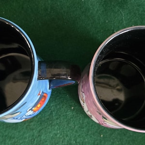 May include: Two ceramic mugs viewed from directly above, both featuring glossy black interiors and black handles. The left mug has a light blue exterior with a partial graphic, while the right mug has a pinkish-purple exterior with a partial graphic.