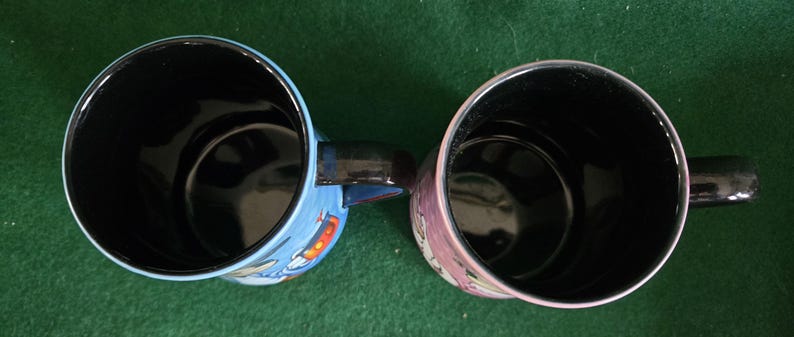 May include: Two ceramic mugs viewed from directly above, both featuring glossy black interiors and black handles. The left mug has a light blue exterior with a partial graphic, while the right mug has a pinkish-purple exterior with a partial graphic.