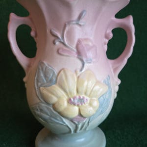 May include: A vintage ceramic vase in soft pastel colors. The vase features a large, embossed flower design in yellow and pink, with blue-green leaves. It has two handles and a pedestal base, perfect for floral arrangements.