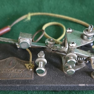 Könnte beinhalten: Ein alter Vibroplex-Telegrafenschlüssel mit einer schwarzen, strukturierten Basis, einer Metallplatte mit der Aufschrift Vibroplex, einem silberfarbenen mechanischen Arm, verstellbaren Schrauben und einem schwarzen Knopf. Ein beiges stoffummanteltes Kabel mit braunem Stecker ist befestigt.