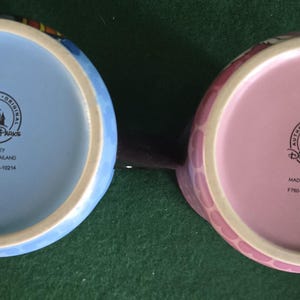 May include: Two ceramic mugs viewed from the bottom, one with a light blue base and the other with a light pink base. Both feature a black circular stamp with a castle icon, the text Disney Parks, Authentic Original, Disney, Made in Thailand.