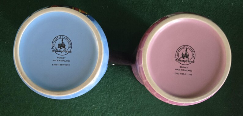 May include: Two ceramic mugs viewed from the bottom, one with a light blue base and the other with a light pink base. Both feature a black circular stamp with a castle icon, the text Disney Parks, Authentic Original, Disney, Made in Thailand.