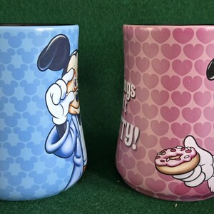 May include: Two ceramic mugs featuring Disney characters. The left mug is light blue with a repeating Mickey Mouse head pattern and a graphic of Mickey. The right mug is pink with a repeating heart pattern and a graphic of Minnie holding a donut.