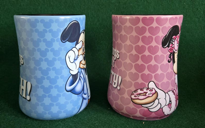 May include: Two ceramic mugs featuring Disney characters. The left mug is light blue with a repeating Mickey Mouse head pattern and a graphic of Mickey. The right mug is pink with a repeating heart pattern and a graphic of Minnie holding a donut.