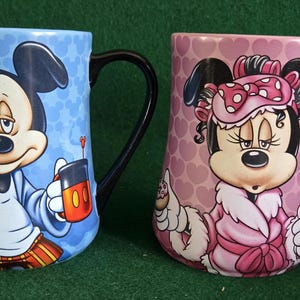 May include: Two ceramic mugs featuring Disney characters. The left mug is light blue with a pattern of Mickey Mouse heads, depicting Mickey in pajamas holding a mug. The right mug is pink with a heart pattern, showing Minnie Mouse in a robe.