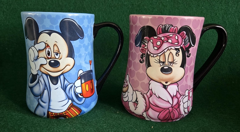 May include: Two ceramic mugs featuring Disney characters. The left mug is light blue with a pattern of Mickey Mouse heads, depicting Mickey in pajamas holding a mug. The right mug is pink with a heart pattern, showing Minnie Mouse in a robe.