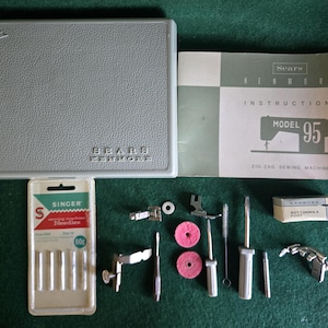 Pode incluir: Um estojo de acessórios para máquina de costura Sears Kenmore cinza, um manual de instruções para o modelo 95, um pacote de agulhas Singer, dois discos de feltro rosa, uma escova pequena, duas chaves de fenda, um pino e vários calcadores.