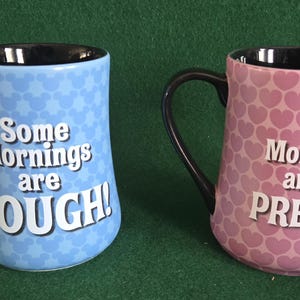 May include: Two ceramic mugs with black interiors and handles. The left mug is light blue with a repeating Mickey Mouse head pattern and the white text Some Mornings are ROUGH!. The right mug is pink with a repeating heart pattern and the white text Mornings aren't PRETTY!.