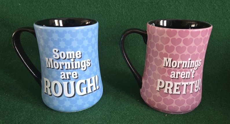 May include: Two ceramic mugs with black interiors and handles. The left mug is light blue with a repeating Mickey Mouse head pattern and the white text Some Mornings are ROUGH!. The right mug is pink with a repeating heart pattern and the white text Mornings aren't PRETTY!.