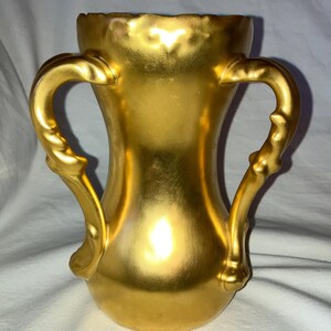 May include: A gold-plated ceramic vase with two handles. The vase has a pearlescent finish and a slightly iridescent sheen.
