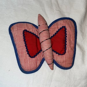 Op de afbeelding: Een handgemaakt decoratief item in de vorm van een vlinder. De vleugels zijn rood en wit gestreept met een blauwe rand. Het midden heeft een rood hartvormig ontwerp. Het lichaam is wit en rood gestreept, omwikkeld met zwart garen.