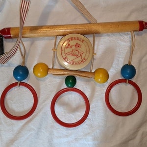 May include: A wooden baby toy with a red, white and blue strap. The toy has a wooden bar with a small drum that says "Chuckle Symphony" on it. The bar has three rings attached to it, each with a different coloured ball. The balls are blue, yellow and green.