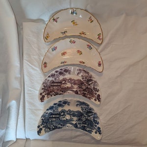 Puede incluir: Cuatro platos de servir vintage en forma de media luna. Los dos platos superiores son blancos con un patrón floral en rosa, amarillo y azul. Los dos platos inferiores son blancos con una escena de paisaje azul y marrón.
