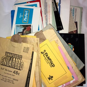 Op de afbeelding: Een stapel vintage papieren enveloppen en flyers voor foto ontwikkeldiensten. De bovenste envelop is bruin met de tekst "Snapshots Enlarged" en "5x7 De Luxe 48c".