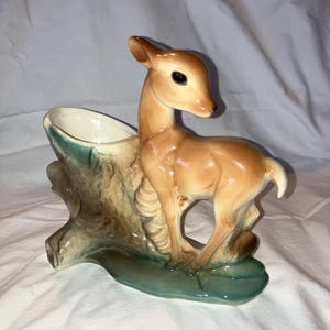 Puede incluir: Una maceta de cerámica con forma de ciervo junto a un tocón de árbol. El ciervo es de color marrón claro con ojos negros y cola blanca. El tocón y la base son marrones y turquesas. La abertura de la maceta es blanca.