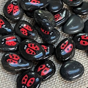May include: A collection of black painted rocks with red ladybug designs, displayed on a burlap bag. A wooden ruler with inch measurements is in the background. The rocks are smooth and glossy.