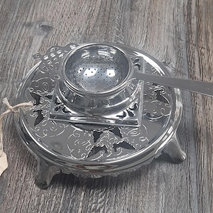 May include: A silver tea infuser set with a decorative stand and a tag that says "Made in England". The infuser has a round strainer and a long handle. The stand has a floral design and three feet.
