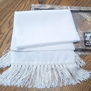 May include: A white scarf with a fringe trim, folded and displayed on a wooden surface. The scarf is made of a smooth fabric and is partially visible inside a clear plastic package with the "Tootal Scarves" logo.