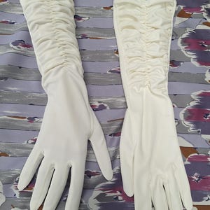 May include: A pair of long, white gloves with a ruched design on the upper arm. The gloves are made of a smooth, opaque fabric and have a classic, elegant style. The gloves are laid out on a patterned surface.