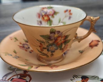 Può includere: Un set da tè con tazza e piattino a fiori con un delicato motivo floreale in tonalità di rosa, viola e verde. La tazza da tè ha un bordo dorato e un manico dorato.