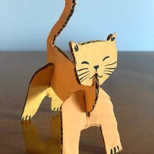 May include: A cardboard cat sculpture painted orange with black details. The cat is standing on all fours with its tail raised in the air.