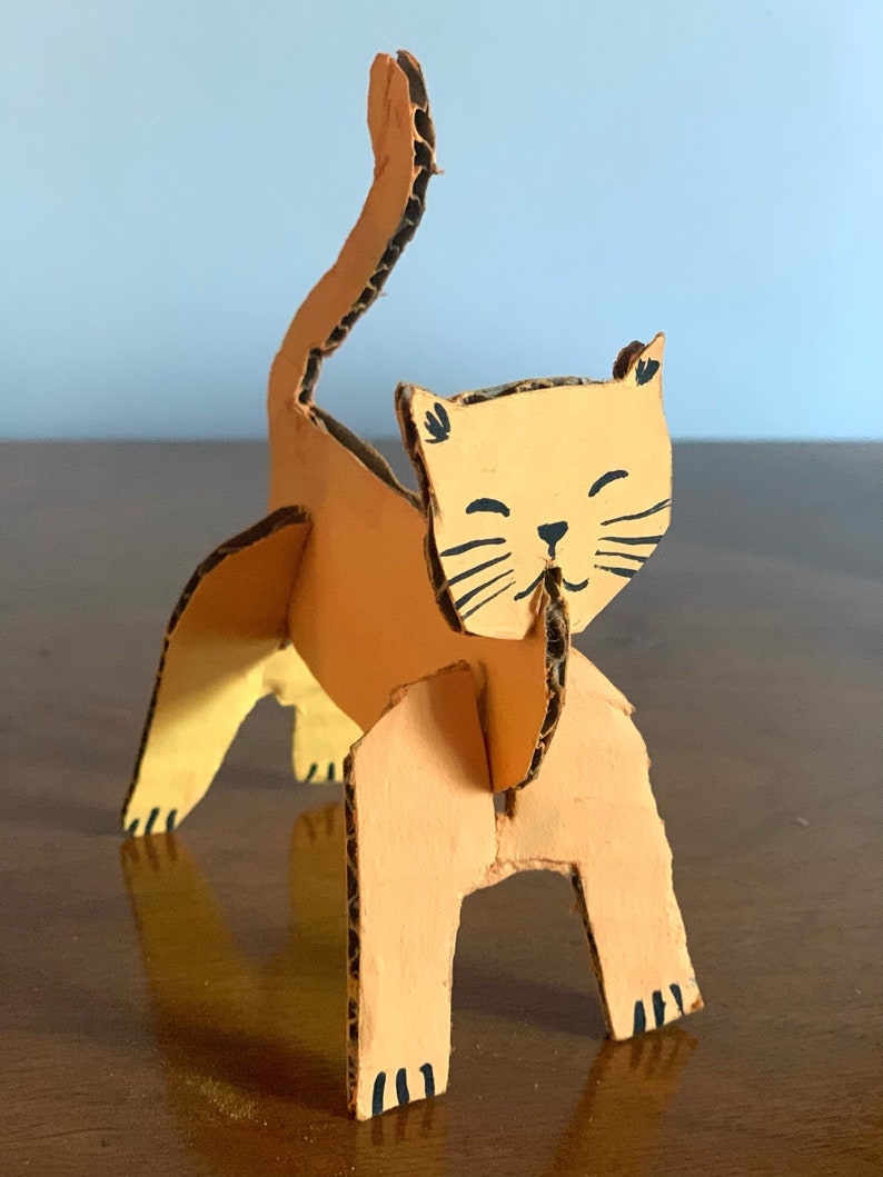 May include: A cardboard cat sculpture painted orange with black details. The cat is standing on all fours with its tail raised in the air.