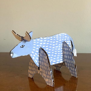 May include: A blue cardboard bull with white polka dots and brown legs with brown polka dots. The bull is standing on a wooden surface.