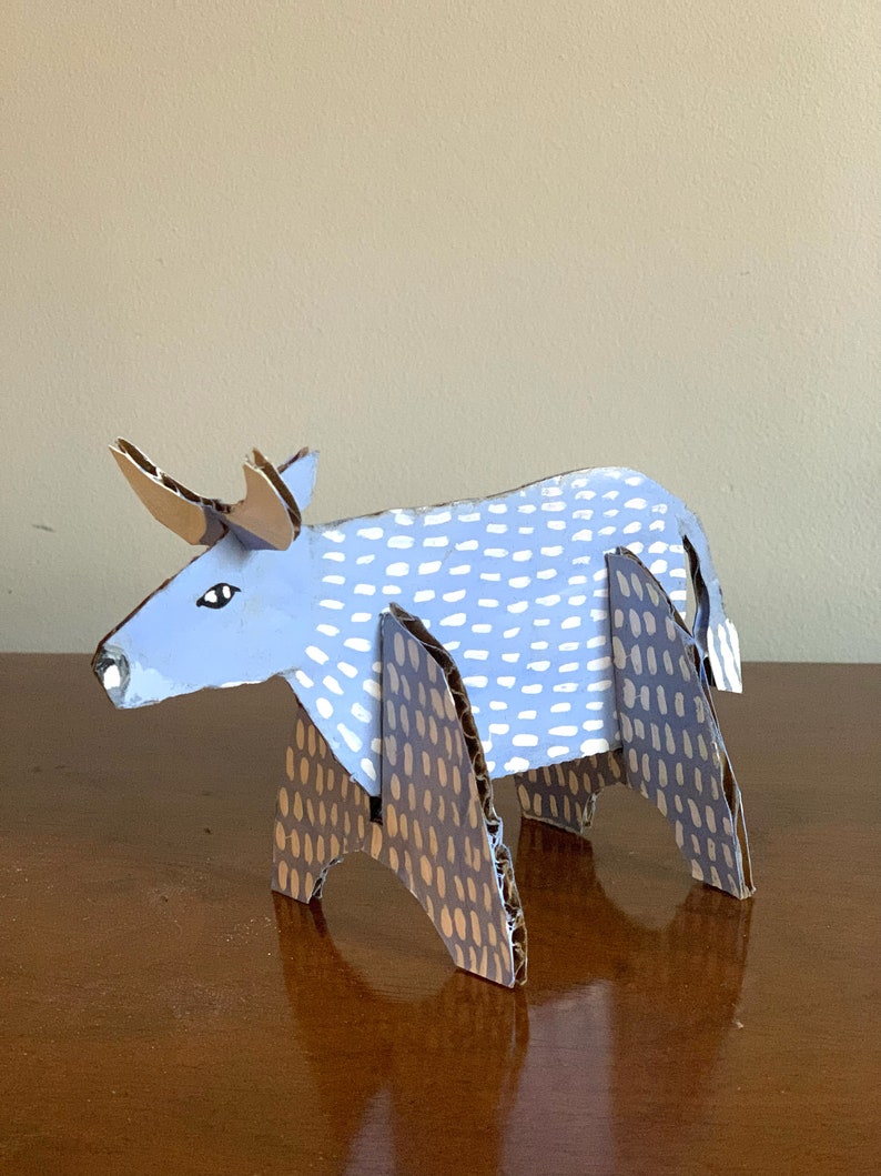May include: A blue cardboard bull with white polka dots and brown legs with brown polka dots. The bull is standing on a wooden surface.