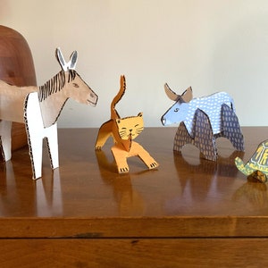 May include: Five cardboard animal figurines, including a donkey, a cat, a bull, and a turtle. The donkey is brown and white, the cat is orange, the bull is blue and white, and the turtle is green and yellow. The figurines are all standing on a wooden surface.