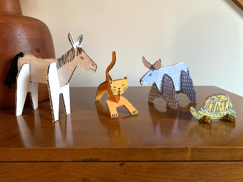 May include: Five cardboard animal figurines, including a donkey, a cat, a bull, and a turtle. The donkey is brown and white, the cat is orange, the bull is blue and white, and the turtle is green and yellow. The figurines are all standing on a wooden surface.