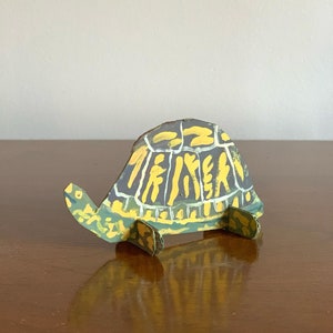 May include: A hand-painted cardboard turtle sculpture with a green, yellow, and black shell. The turtle is standing on a wooden surface.